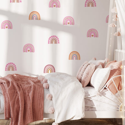 A cozy childrens room features a white bed with a pink and white blanket. My Stores Rainbow Wall Stickers in pastel hues adorn the wall. Soft pillows and a canopy with hanging clouds complete the whimsical decor.