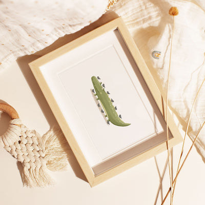 A wooden-framed Safari Animals Nursery Print by My Store, featuring a green crocodile on white, lies on beige. Its surrounded by a white textile with gold dots, a decorative beige woven tassel, and dried wheat stalks for an enchanting safari-themed display.