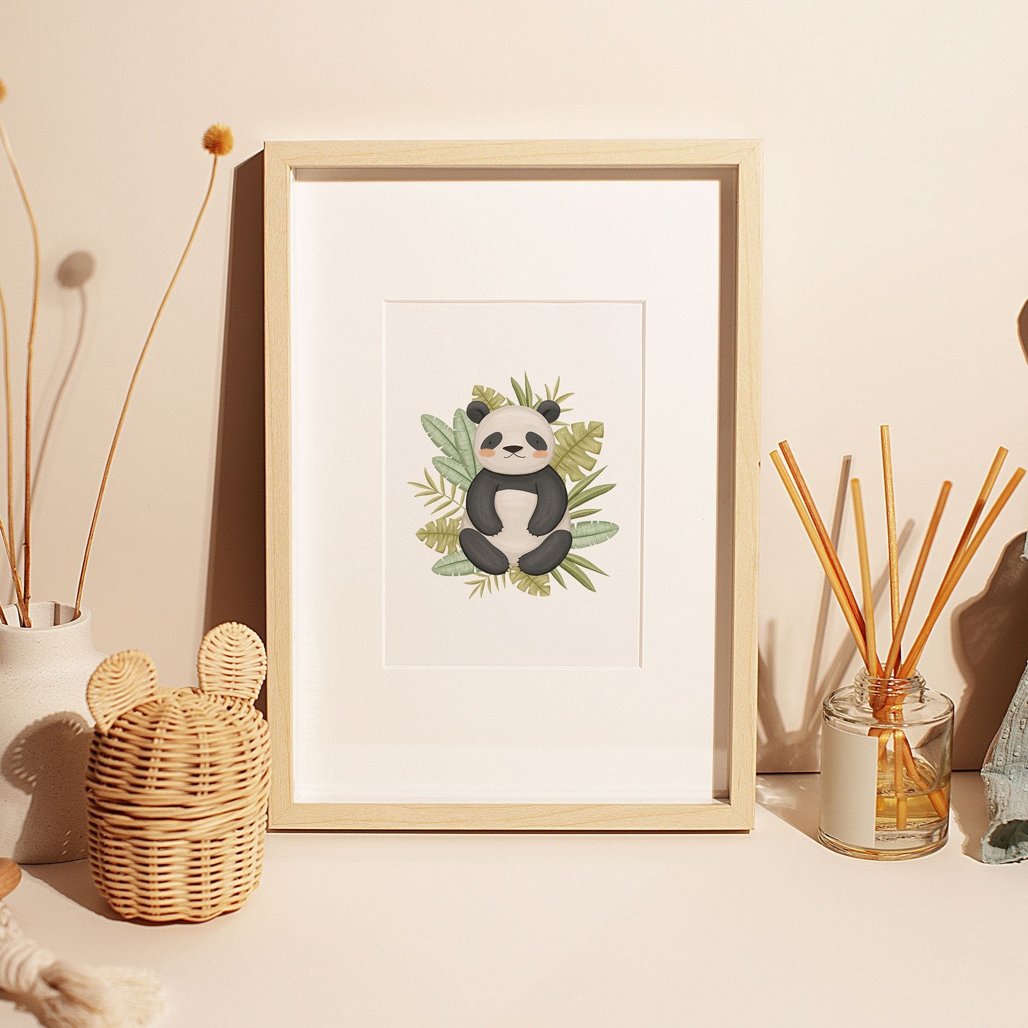 A framed Safari Animals Nursery Prints illustration from My Store, featuring a panda among leaves, sits on a light table. Nearby are decorative items like a wicker basket, dried flowers in a vase, and reed diffusers. This softly lit scene creates a warm ambiance perfect for any nursery.
