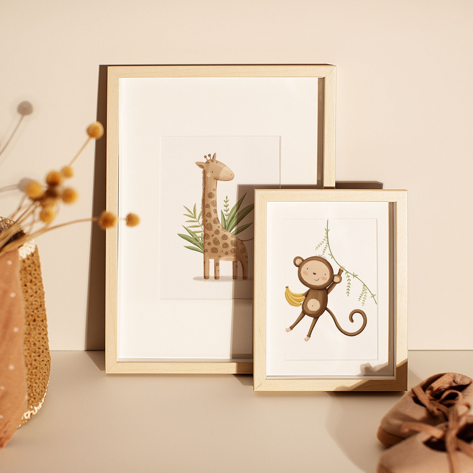 Two of My Stores Safari Animals Nursery Prints sit on a light surface with decor: one features a giraffe with leaves, and the other shows a monkey swinging on a vine with a banana.