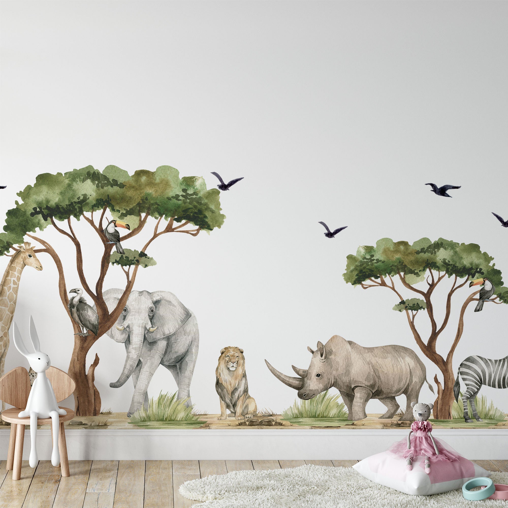 A kids room boasts a vibrant mural of safari animals with Safari Animals Nursery Stickers by My Store—giraffe, elephant, lion, rhinoceros, and zebra under trees. Watercolour decals add charm with flying birds above. A plush rabbit sits on a chair near a doll on a cushion and a toy wagon on the floor.