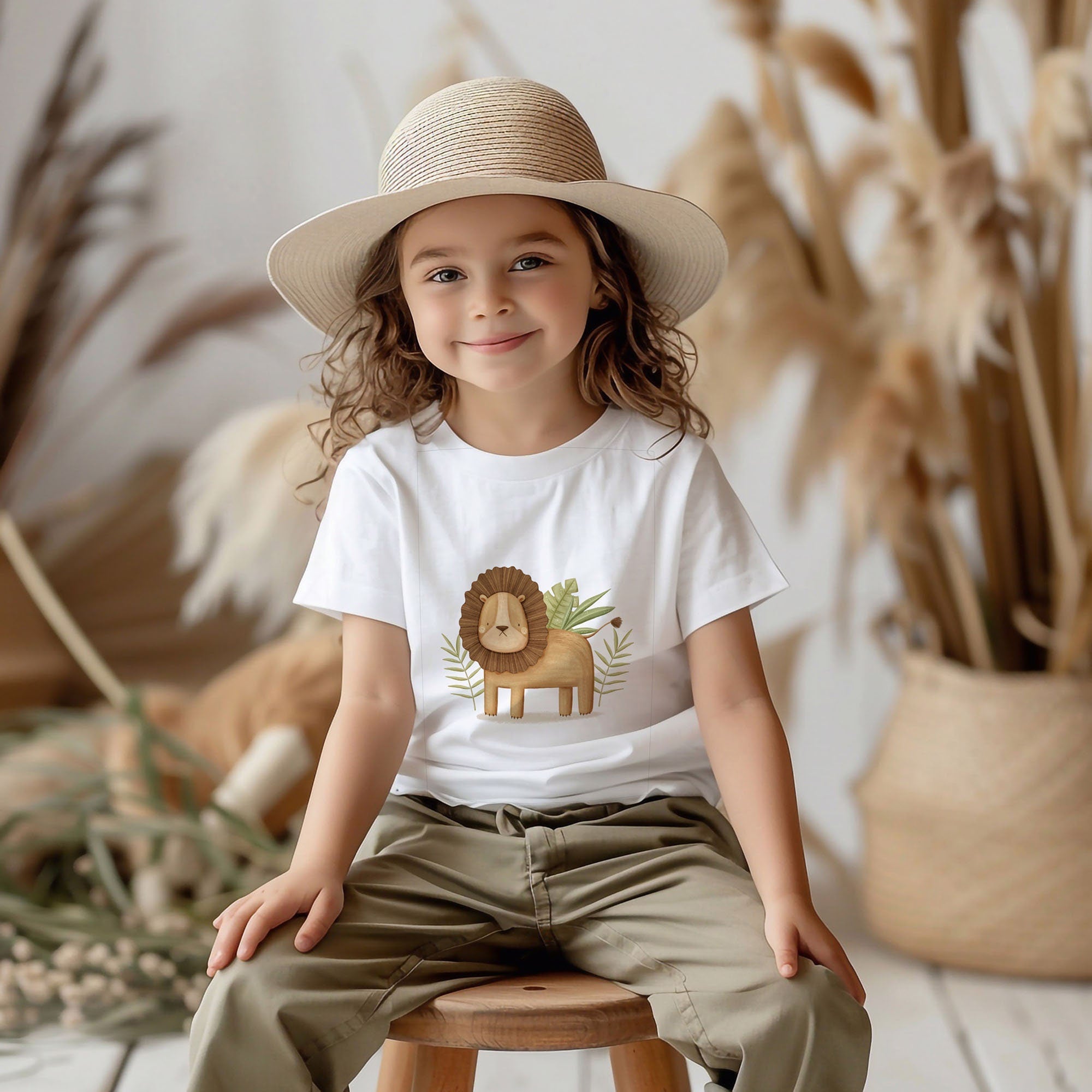 A curly-haired child in a straw hat and My Stores Safari Animals Toddler T Shirt - White sits on a wooden stool. The 100% ringspun combed cotton shirt is machine washable, set against dried plants and neutral tones for a cozy, natural backdrop.
