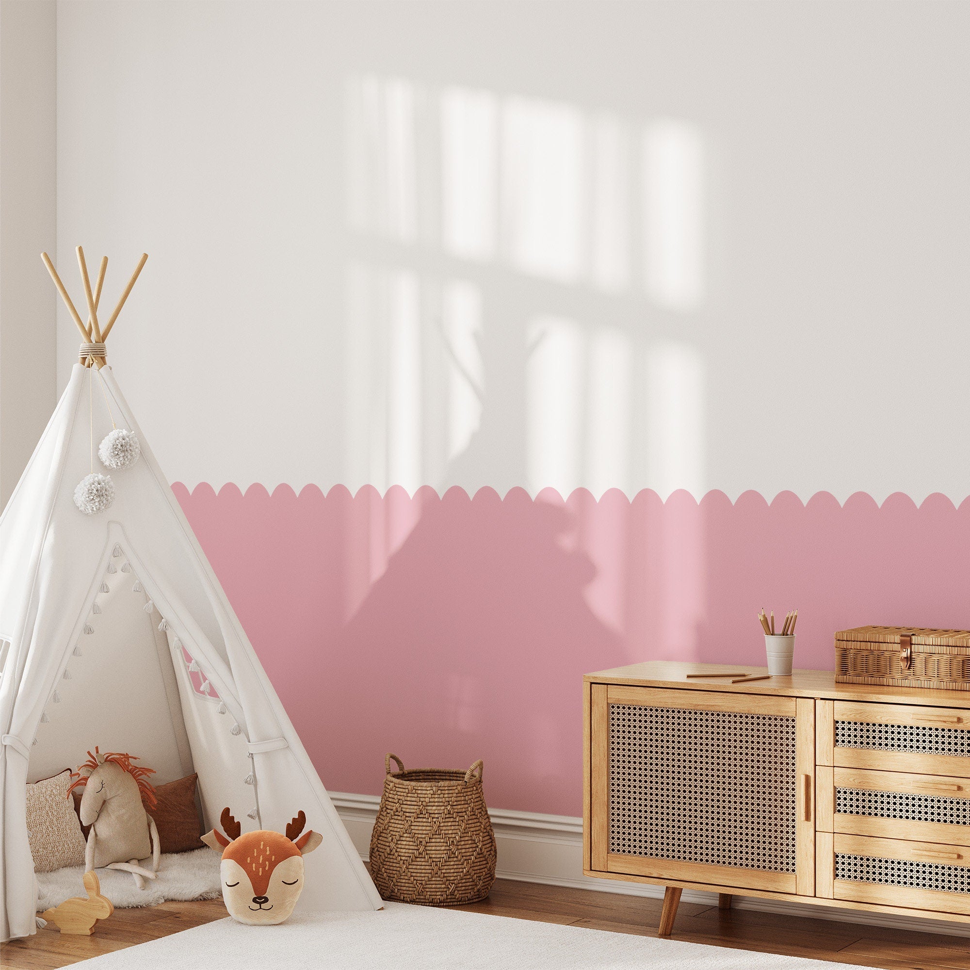 A cozy childrens room features a white and pink wall enhanced with My Stores Scalloped Wall Stencil, a pom-pom teepee, deer pillow, wicker basket, and wooden sideboard. Soft light fills the room, creating a warm and inviting atmosphere.