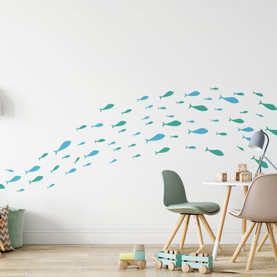 A modern childrens room with wooden floors features white walls adorned with Shoaling Fish Stickers by My Store in teal and blue. The nursery has a small round table, two chairs, a wooden toy car, and a lamp. Wood blocks spell JULK.