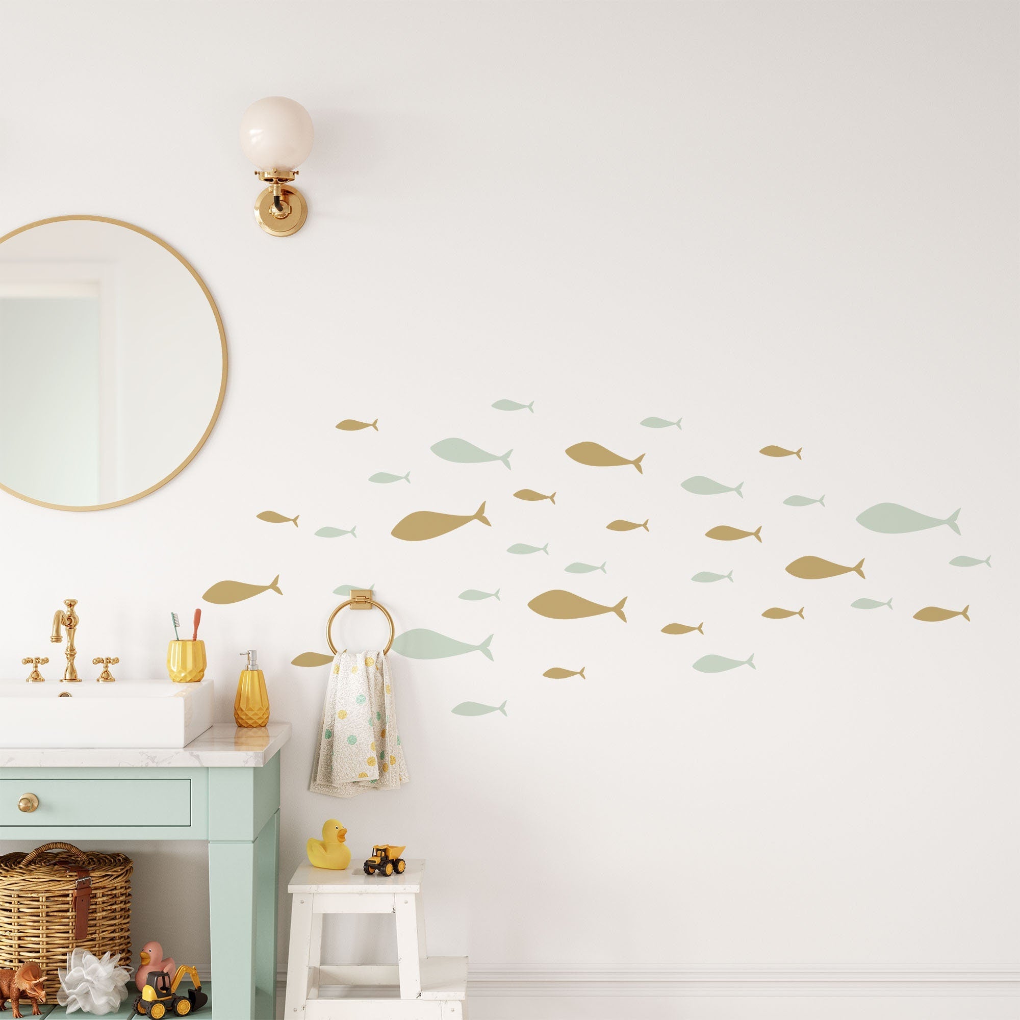 In a bathroom scene, My Stores Shoaling Fish Stickers in gold and light green add nursery decor to the walls. A round mirror hangs above a white sink with a pale green cabinet, topped with toiletries, a duck figurine, and towel. A wall-mounted light fixture is positioned over the mirror.