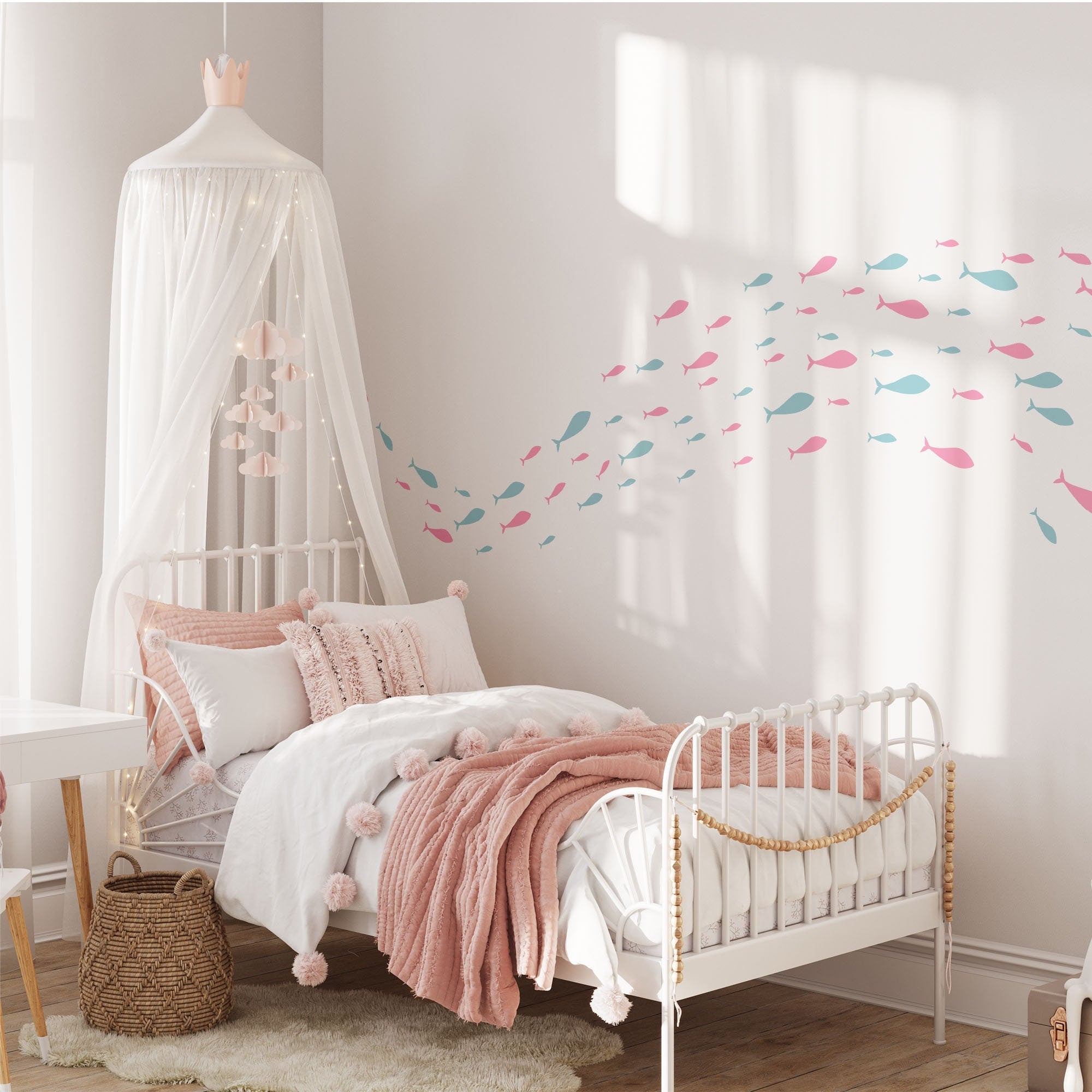 A childrens bedroom with charming nursery decor features a white metal bed with pink and white bedding. A canopy with lights hangs above, enhanced by My Stores Shoaling Fish Stickers in pink and blue. A wicker basket sits on the floor, and a desk is partially visible on the left.