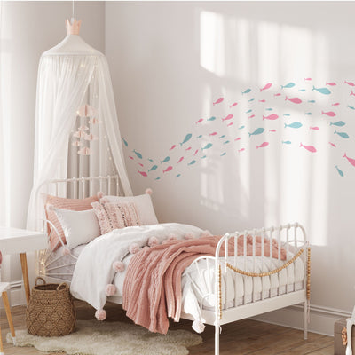 A childrens bedroom with charming nursery decor features a white metal bed with pink and white bedding. A canopy with lights hangs above, enhanced by My Stores Shoaling Fish Stickers in pink and blue. A wicker basket sits on the floor, and a desk is partially visible on the left.