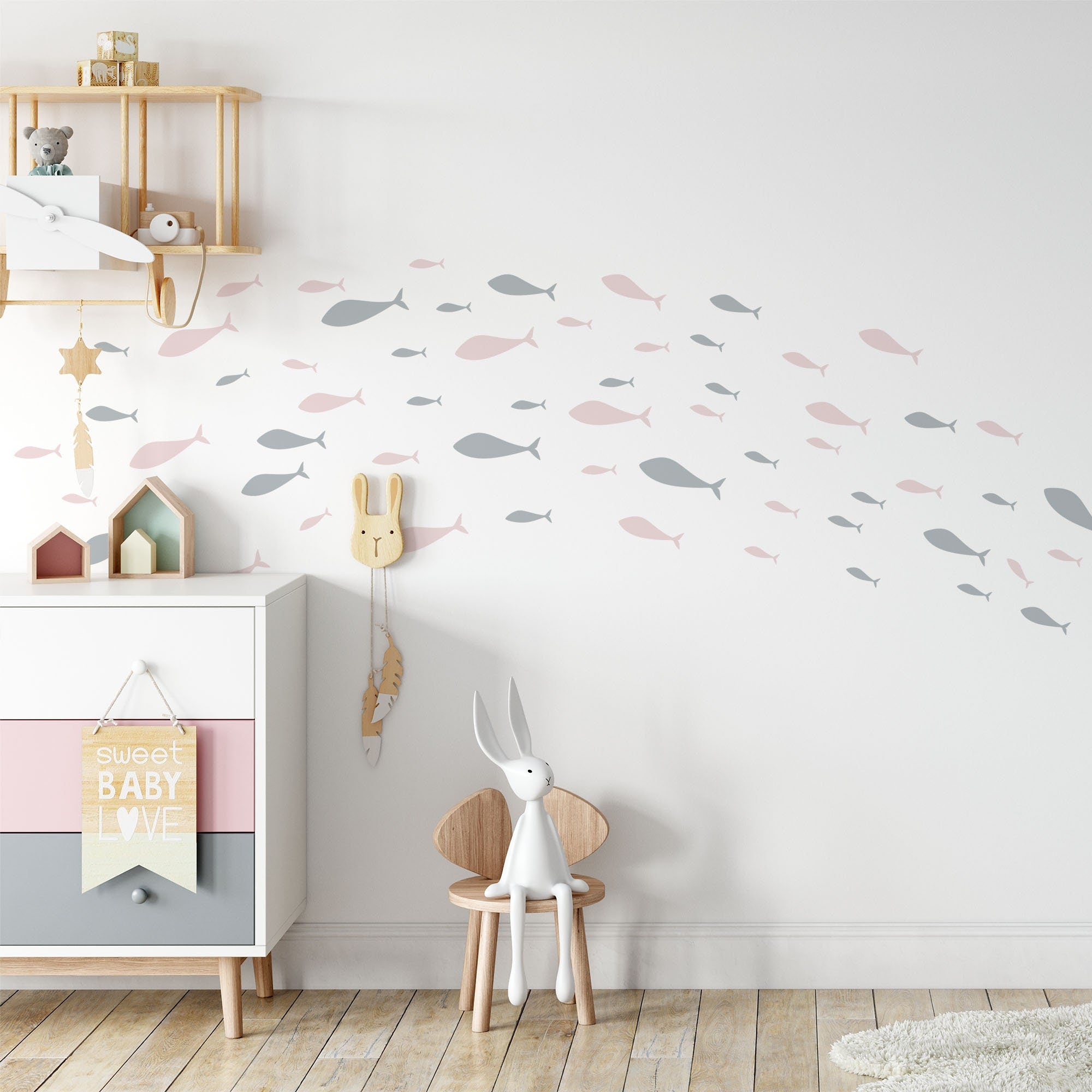 A nursery with white walls is adorned with My Stores Shoaling Fish Stickers in gray and pink. A white dresser boasts a Sweet Baby Love sign among charming decor, while a small table holds a plush bunny and the floor features a cozy rug.