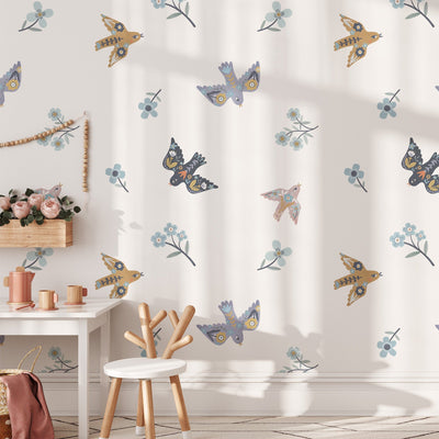 The serene childrens room features white walls decorated with My Stores Summer Mood Wall Stickers, showcasing vibrant bird and floral patterns. A wooden chair and table sit next to a plush toy-filled shelf, adding a cozy touch to the playful nursery setup.