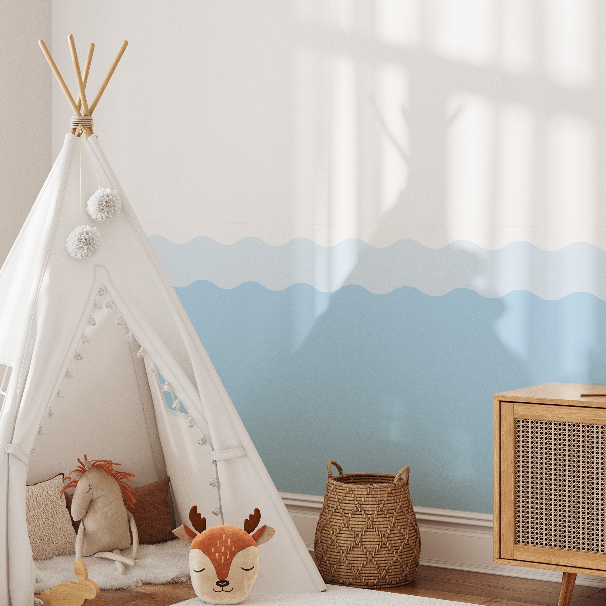 A cozy childrens playroom includes a white teepee tent with fluffy pom-poms, a stuffed deer, and lion toy inside. The walls use My Stores Wave Wall Stencil for the soft blue wave design. Nearby are a wicker basket and wooden cabinet, while sunlight plays on the wall.