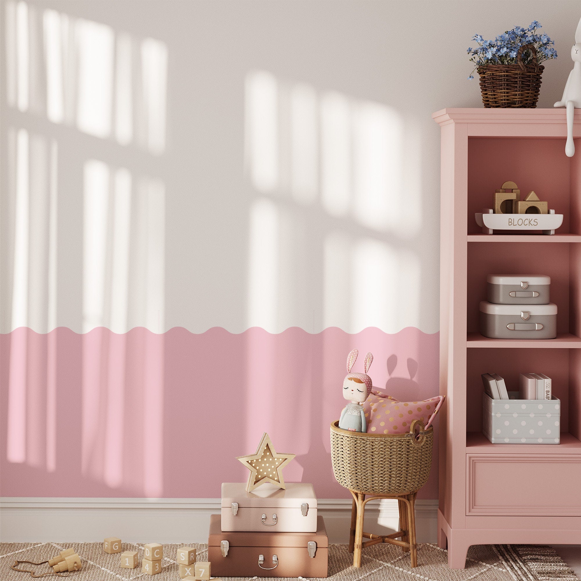 A cozy childrens room features pink and white walls with My Stores Wave Wall Stencil. A pink shelf holds toys and books, a wicker basket with a doll is beside suitcases, and a star-shaped lamp. Soft sunlight through the window casts playful shadows on the floor.