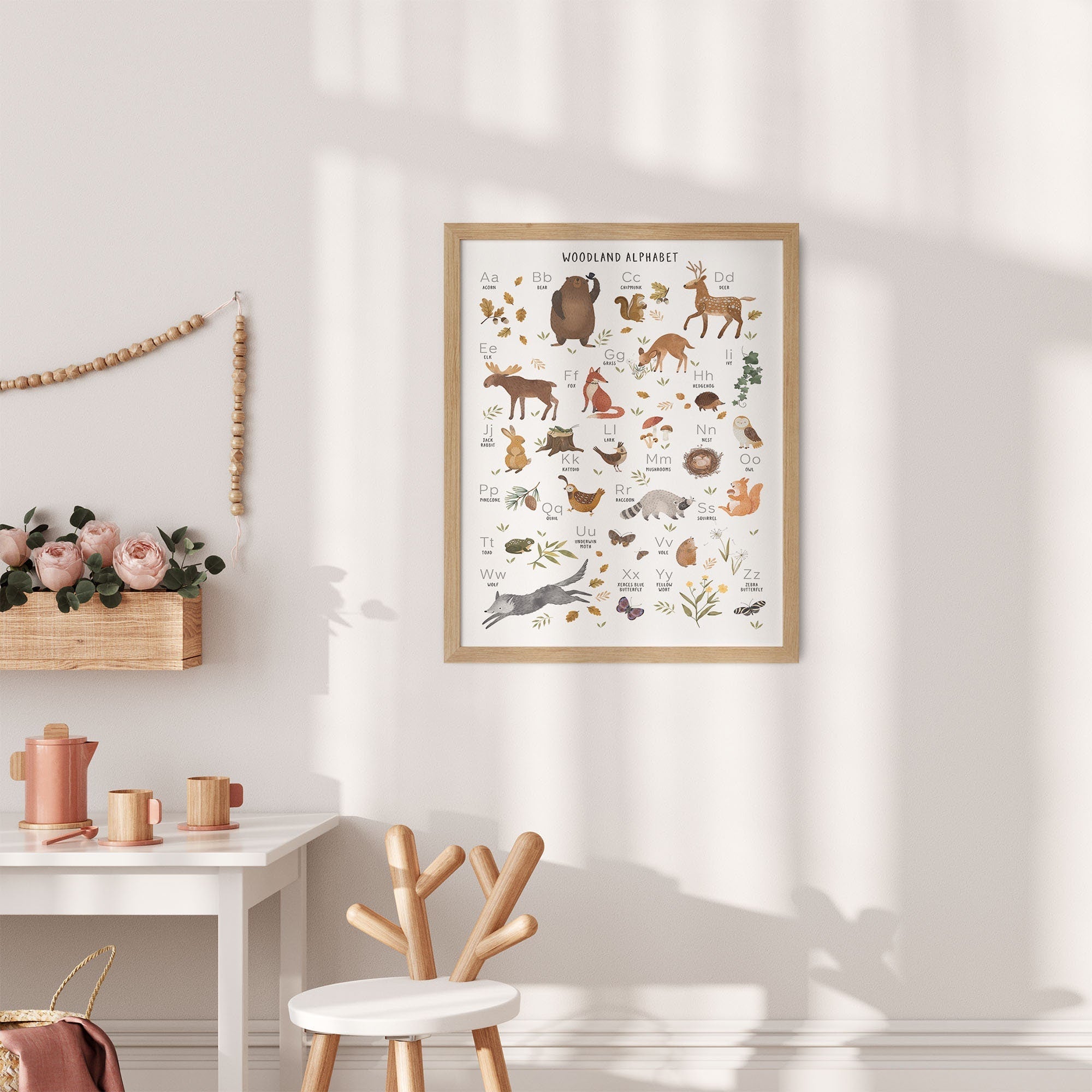 A cozy room showcases the Woodland Alphabet Nursery Print from My Store on a light wall, enhancing educational value. A white table with a wooden chair holds pink flowers in a basket, along with a teapot and cups. Sunlight casts soft shadows for a warm atmosphere.