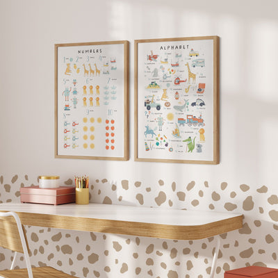 A childrens room features a wooden desk, white chair, and My Stores Numbers & Alphabet Nursery Print. One poster displays numbers with illustrations, the other showcases the alphabet with images. Warm lighting and a spot-patterned wall complete the playful decor.