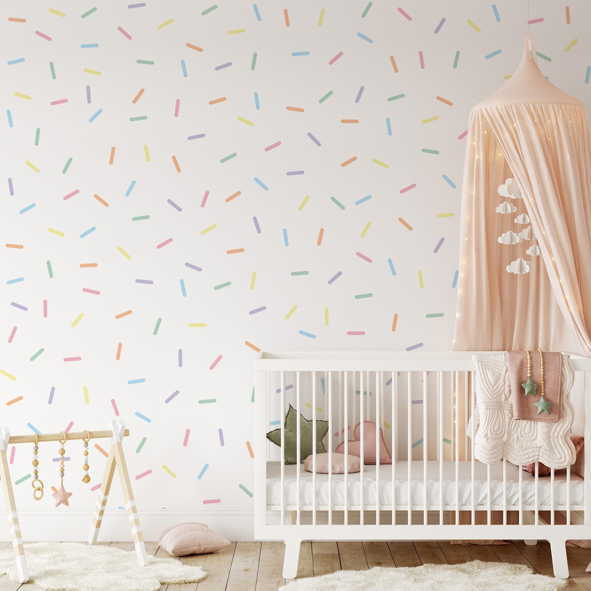 A nursery with a white crib with pink bedding, a hanging peach canopy adorned with star lights and cloud cutouts, and a wooden baby gym. The wall is decorated using My Stores Pastel Sprinkle Stickers as removable nursery decor.