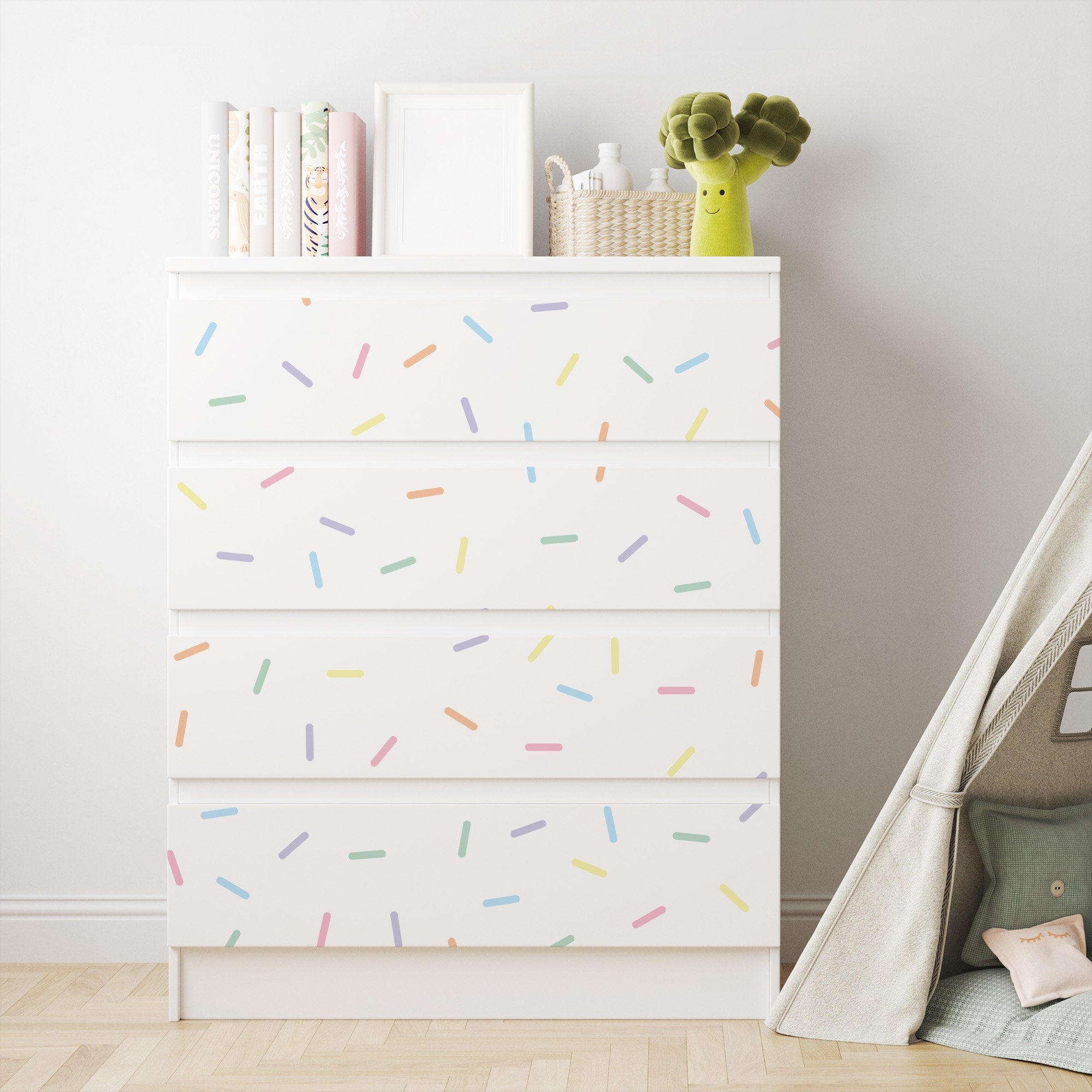 A white dresser, enhanced with Pastel Sprinkle Stickers by My Store, stands next to a small fabric play tent. The minimalist setup is adorned with nursery decor charm: books, a blank picture frame, a woven basket, and a plush broccoli toy top the playful scene.