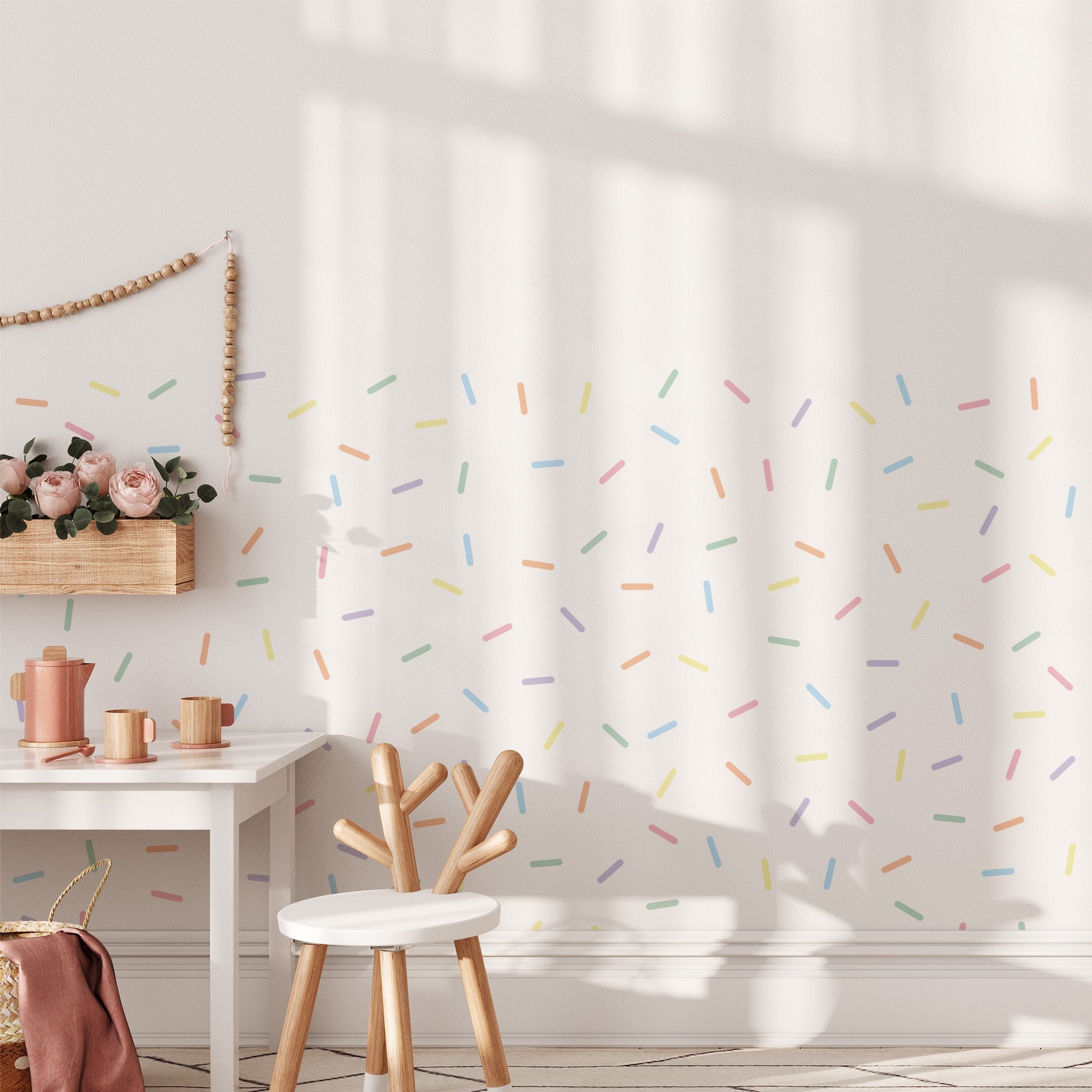 A bright, modern room showcases a minimalistic white desk and chair. The wall features colorful Pastel Sprinkle Stickers from My Store, adding a playful touch. A small shelf with pink flowers complements the scene as sunlight casts soft shadows on the floor and wall.