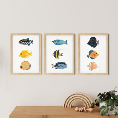 Displayed on a light wall, the Reef Fish Nursery Print by My Store showcases three framed illustrations of colorful reef fish. Beneath them sits a wooden tabletop adorned with a decorative arch stacker and a small green plant, offering nursery art charm and educational fun.