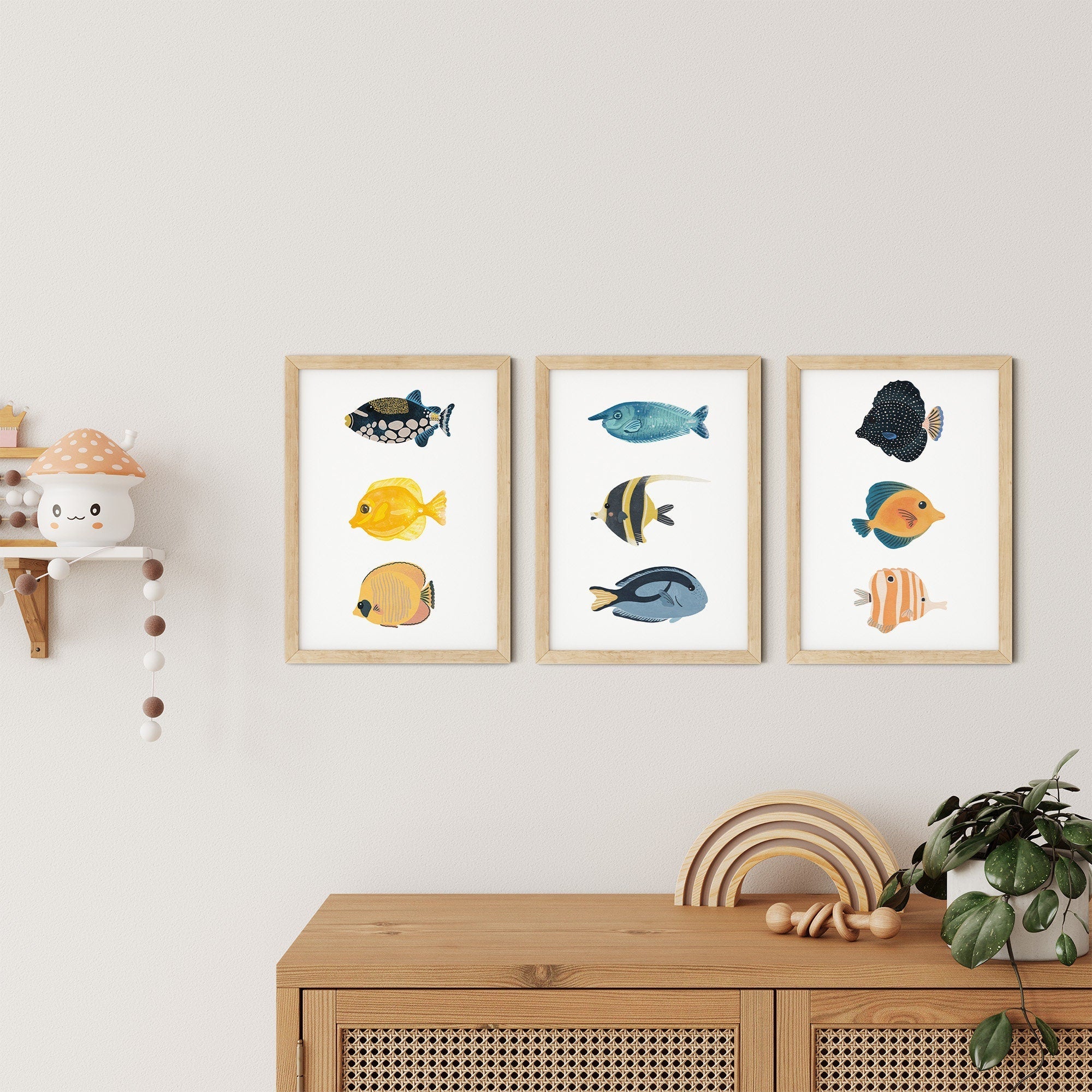 Three My Store Reef Fish Nursery Prints hang on a light wall above a wooden cabinet. To the left, a small decorative shelf holds a mushroom item. A plant atop the cabinet adds greenery to this charming nursery display.