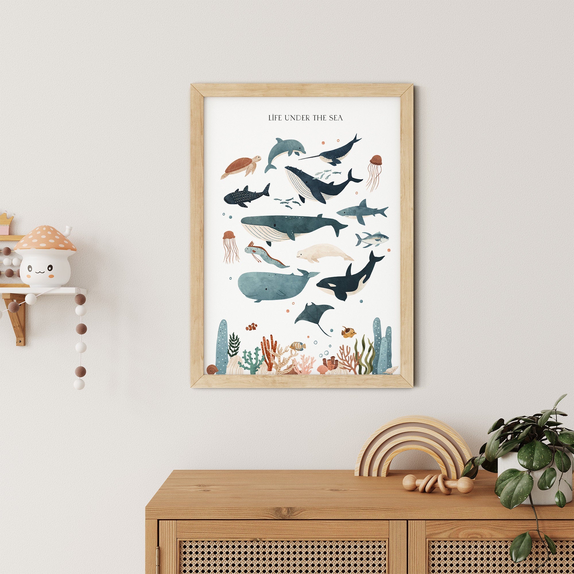 A framed Sea Animals Nursery Print by My Store, featuring ocean creatures like whales, sharks, and jellyfish, decorates a beige wall above a wooden cabinet adorned with plants and a shelf featuring a smiling mushroom lamp.