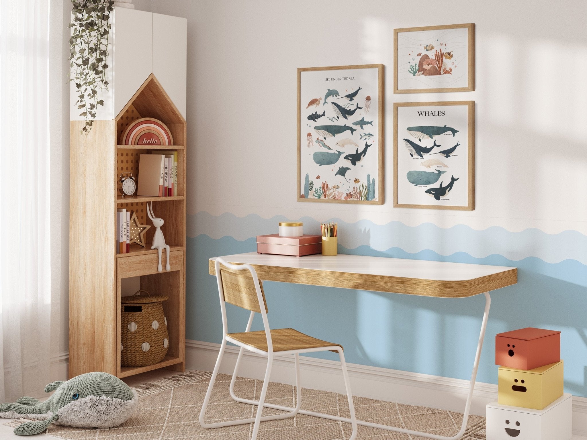 A cozy childrens room showcases a wooden desk, chair, and bookshelf with My Stores Sea Animals Nursery Print. Above the desk is whale-themed wall art, complemented by a green whale plush toy on the rug. The blue wave walls and colorful storage boxes create a playful sea adventure ambiance.
